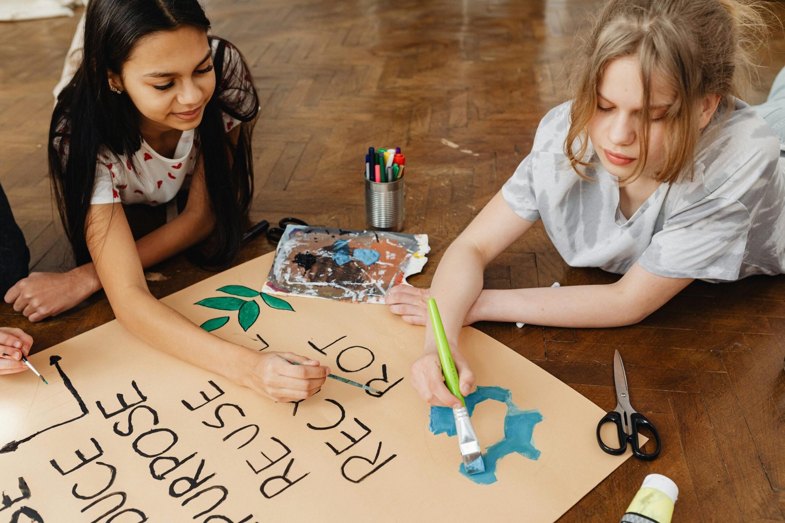 Two teenage girls painting a poster for an art project, promoting recycling and creativity. Indoors setting.